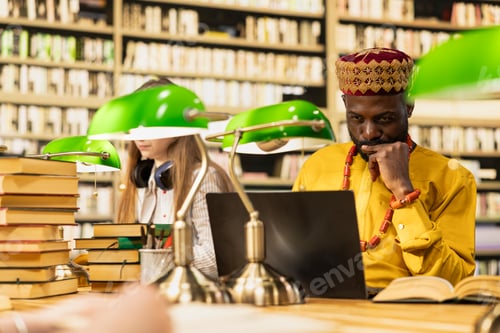 Preview: Adult learner in african traditional robe works on laptop at library table