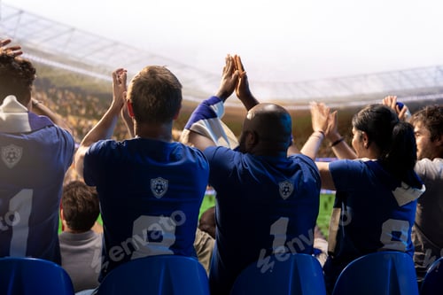 Preview: Rear view of football supporters inside packed stadium clapping during game