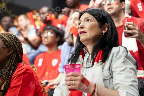 Preview: Mature female football fan with red scarf and pink cup watching tense match
