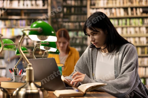 Preview: Focused student using laptop for note taking beside open book at library