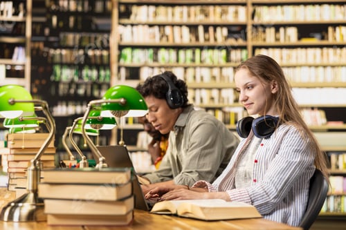 Preview: Young woman with headphones typing on laptop near stacked books
