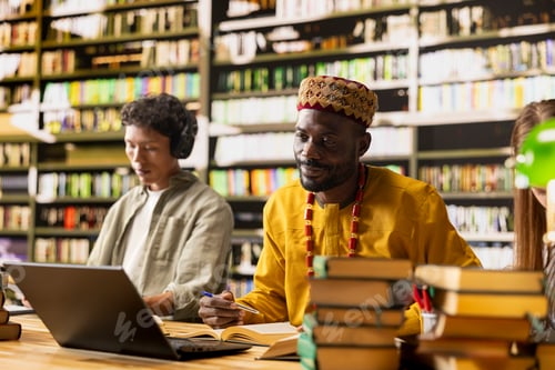 Preview: Black man in african traditional clothing studying with open book and laptop