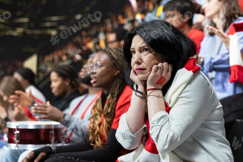 Preview: Mature female football supporter covering ears in loud stadium crowd