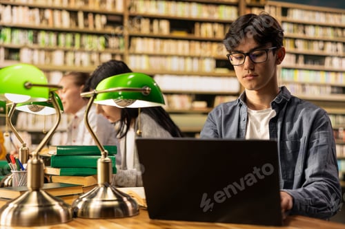 Preview: Asian student with glasses working on laptop at university library table
