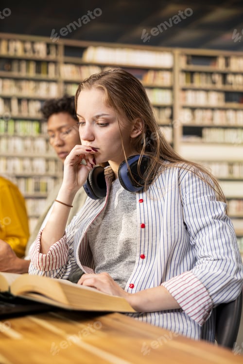 Preview: Young adult learner reading open book beside peers at wooden library desk