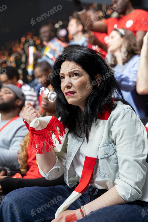 Preview: Concerned mature female football supporter sitting in stadium crowd