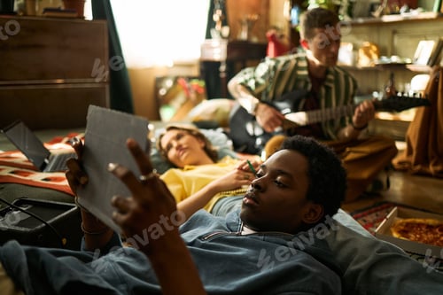 Preview: Black Man Relaxing with Tablet While Friends Playing Guitar