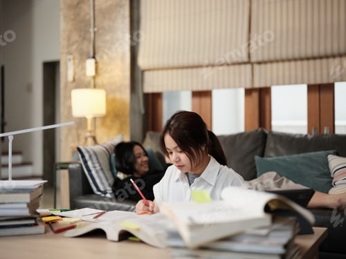 Preview: Asian teenage girl learning at home, her friend relaxing on sofa.