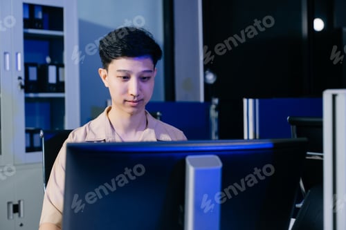 Preview: Happy Asian young man smiling while typing on his laptop at a modern corporate office desk at night.