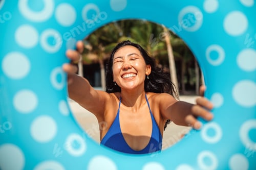 Preview: Cheerful woman posing with a blue swim ring on a sunny tropical beach