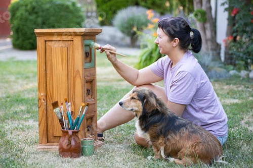 Preview: Woman painting a wooden furniture while sitting with her dog in the garden. DIY concept.
