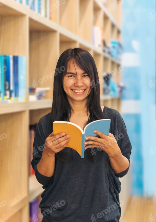 Preview: Asian woman holding book reading at bookshelf in university library. Young woman relaxation read ope