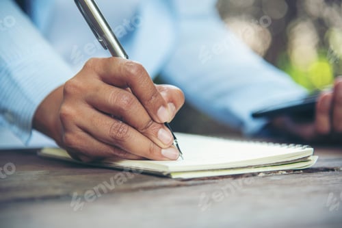 Preview: Man hands writing notebook diary with coffee cup and smartphone on wood desk. Close up man hands usi