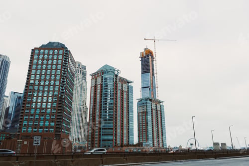 Preview: Tall buildings under construction in a city skyline on a cloudy day near the road with traffic