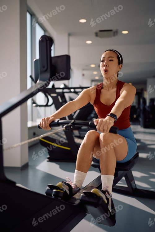 Preview: Female athlete practicing on rowing machine in health club.