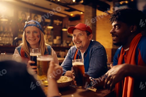 Preview: Happy diverse sports fans toasting with beer while watching sports match in a bar.