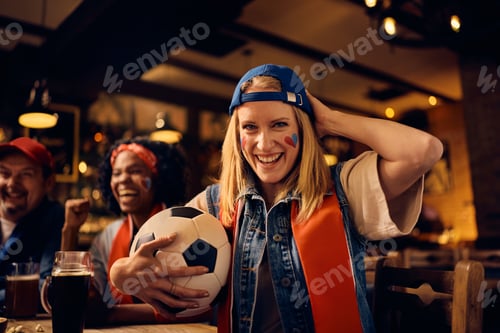 Preview: Happy female fan watching soccer match with friends in a pub and looking at camera.