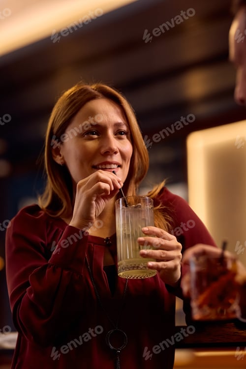 Preview: Smiling woman drinking cocktail while talking to a friend at bar counter.