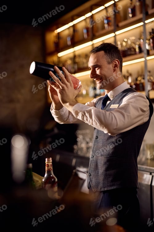 Preview: Happy bartender using cocktail shaker while making a drink at bar counter.