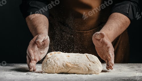 Preview: Professional baker shaping fresh dough with flying flour on dark background for artisan bakery adver