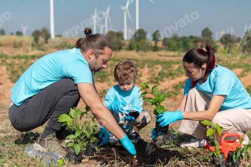 Preview: Volunteer Family Team Planting Trees and Seedlings Together in Open Field with Wind Turbines in Back