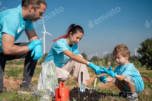 Preview: Volunteer Family Team Planting Trees and Seedlings Together in Open Field with Wind Turbines in Back
