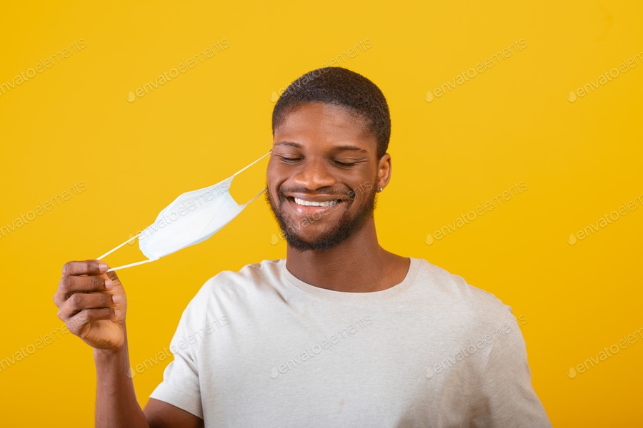 Cover for End of covid-19 pandemic. Happy black man taking off protective face mask, standing over yellow